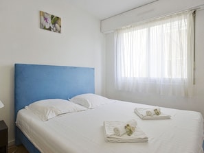 Uma cama bem arrumada com lençóis brancos e cabeceira azul, uma janela com cortinas finas e um pequeno quadro na parede.