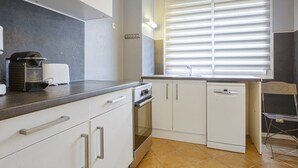 A modern kitchen with white cabinets, a built-in oven, and a window with blinds. - Appartement Herta - Welkeys (Biarritz)