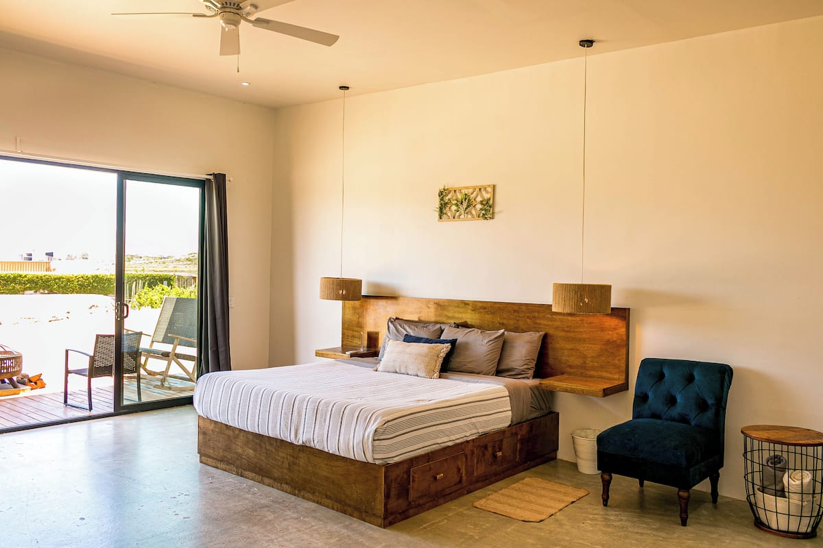 Quarto com cama grande, cabeceira de madeira, mesa de apoio, uma cadeira e vista através de uma porta de vidro.