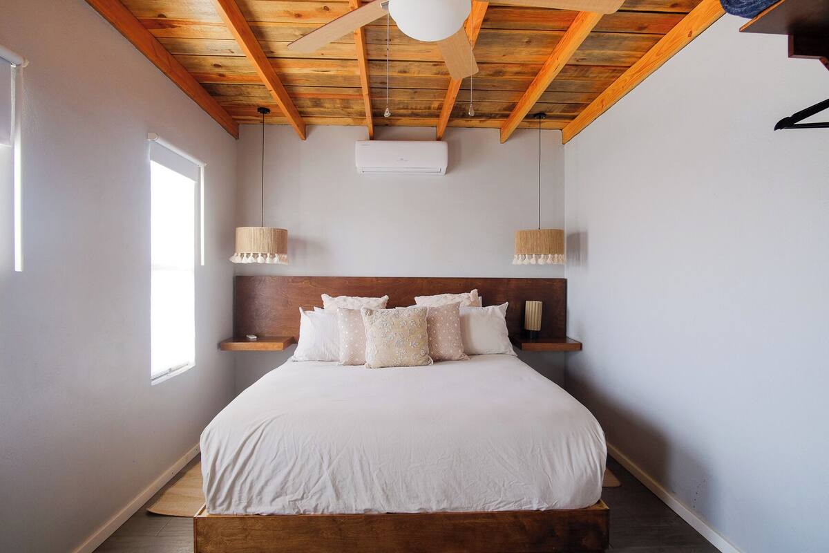 Quarto com teto de madeira, cama com roupa de cama branca, duas luminárias de cabeceira e uma janela.