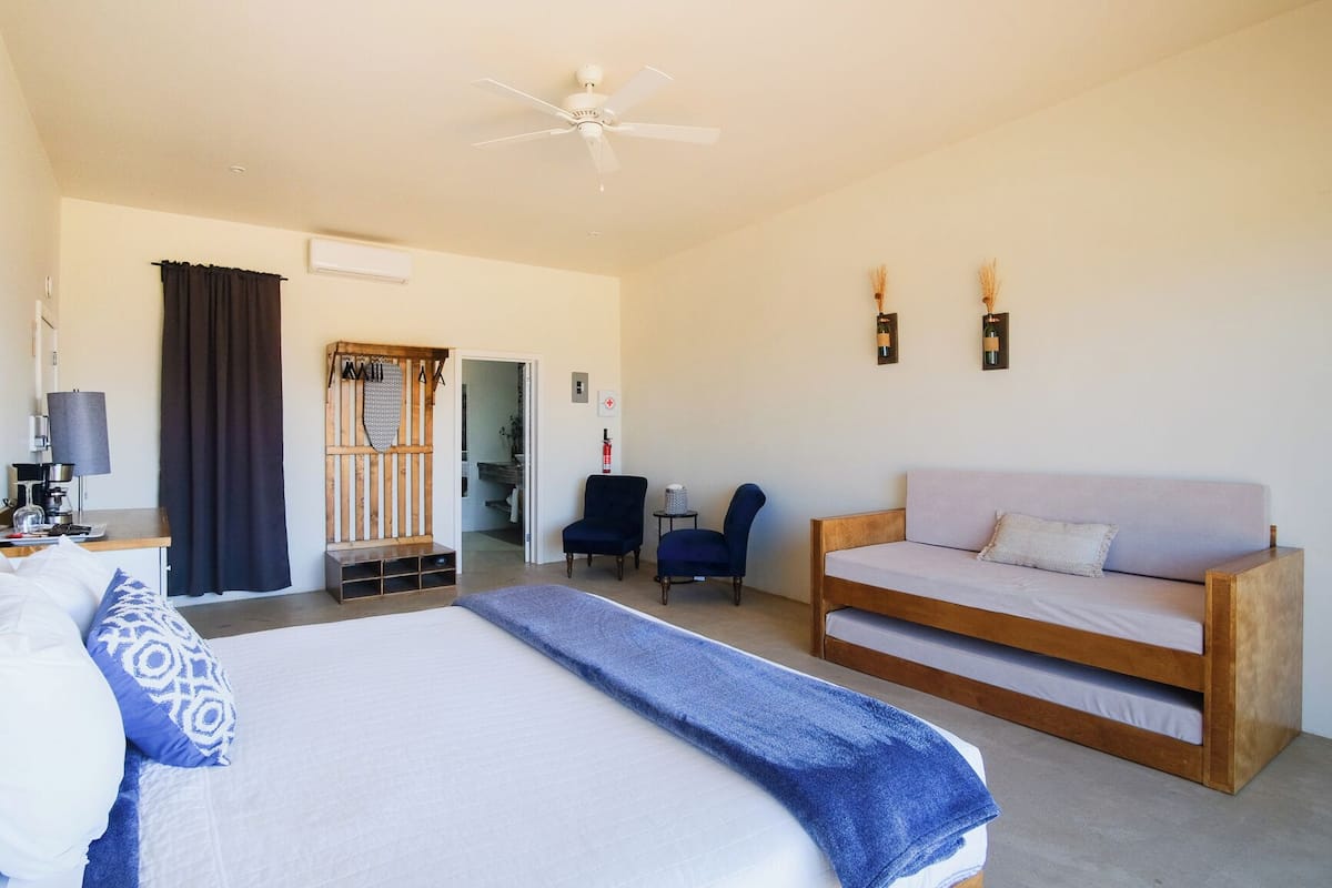 Quarto de hotel com cama, sofá, escrivaninha com luminária, cadeira, ventilador de teto e porta que dá para outro cômodo.