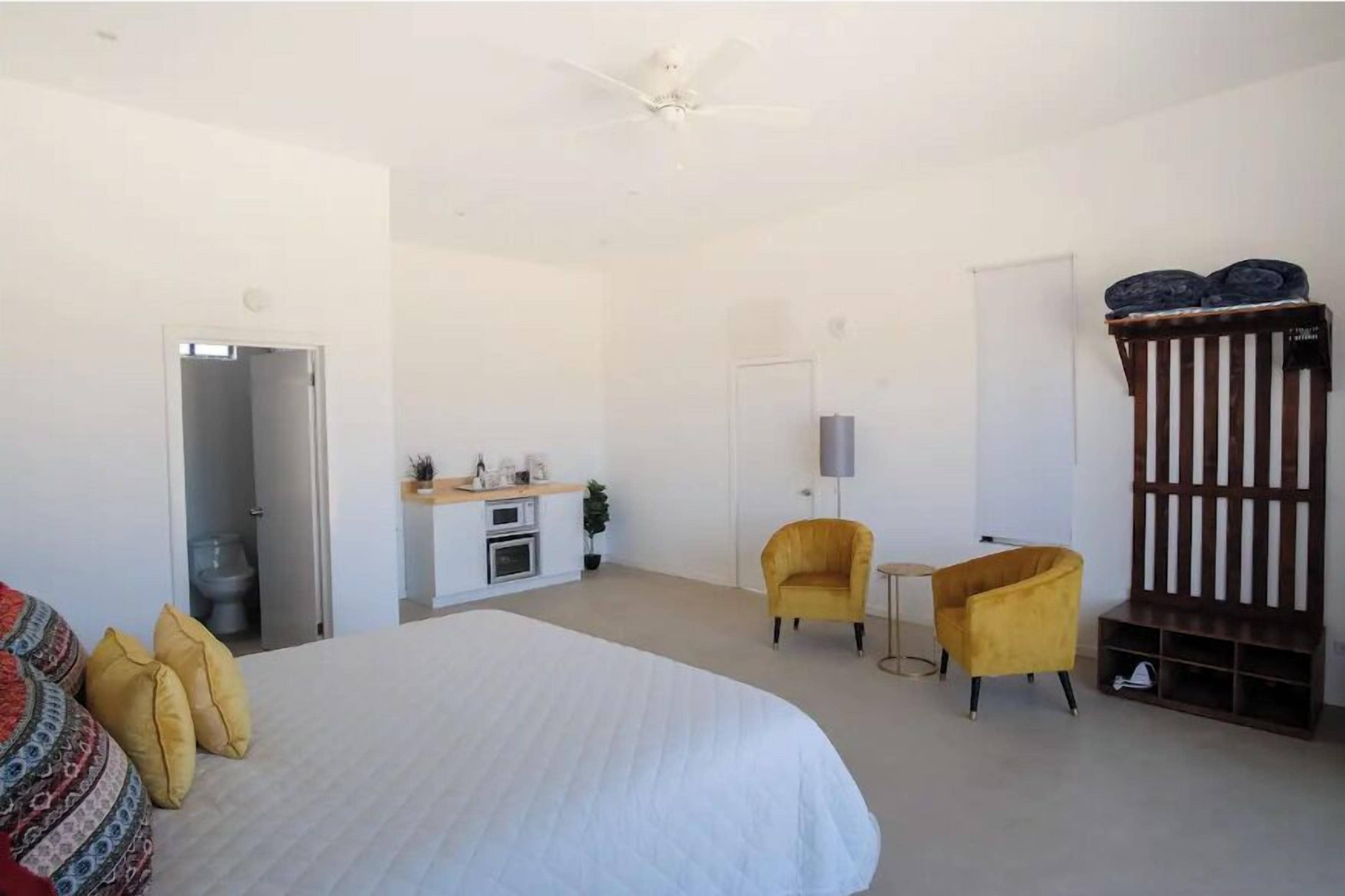 A bedroom with a large bed, a small kitchenette, two yellow armchairs, and a wooden bench.