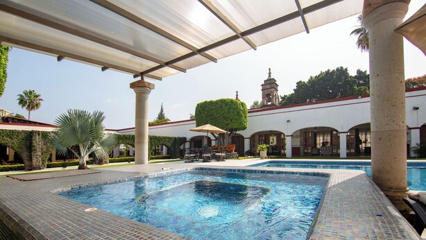 Uma piscina com bordas azulejadas, rodeada por palmeiras e uma área coberta com colunas.
