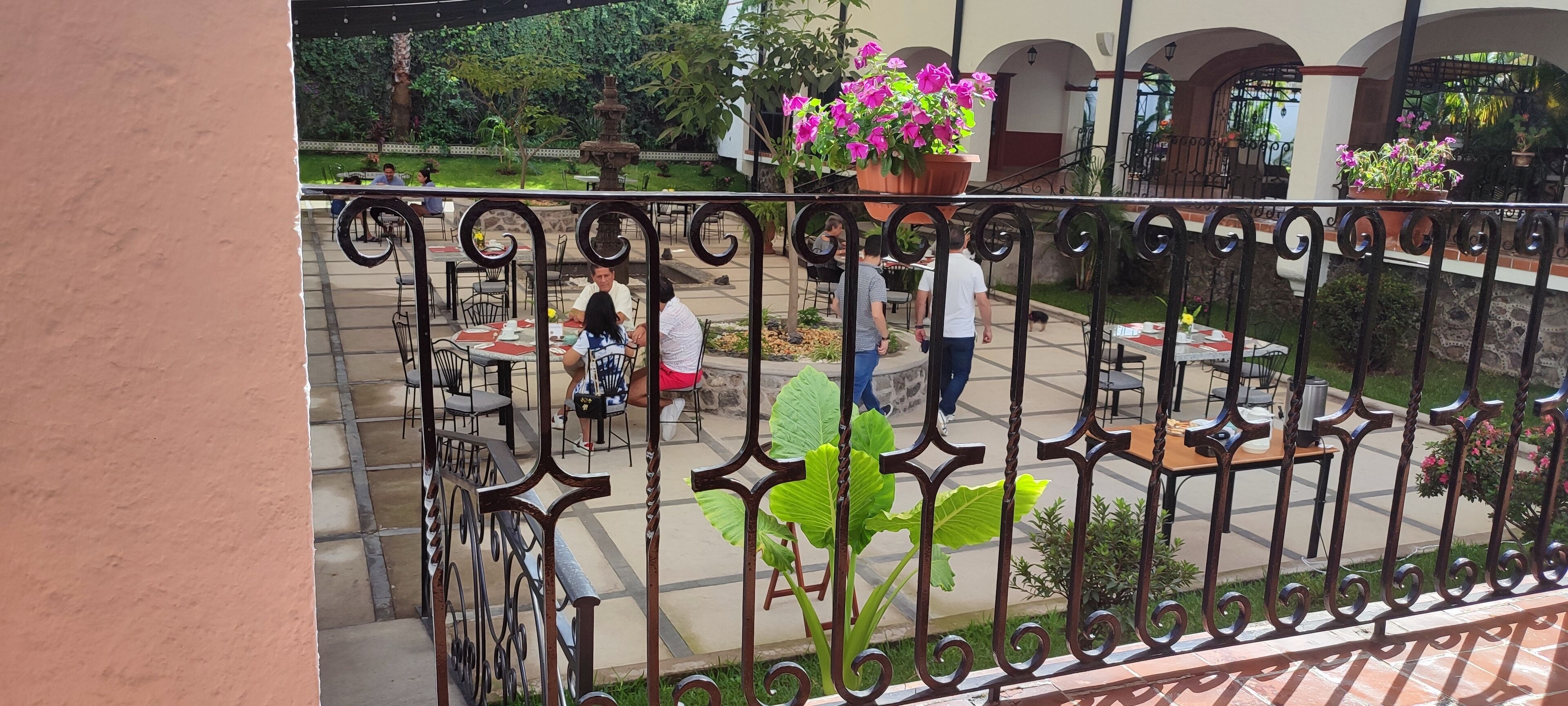 Un área de patio al aire libre con mesas y sillas, rodeada de macetas con plantas y una fuente.