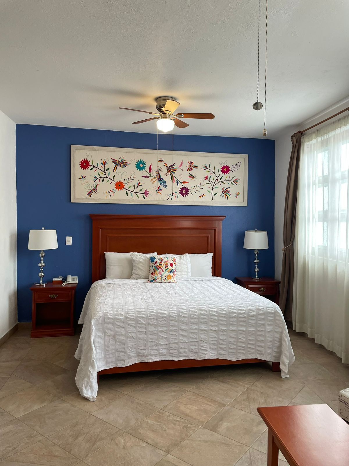 A hotel room with a bed, a ceiling fan, a nightstand with a lamp, a bedside table with a lamp, and a large floral artwork on the wall.