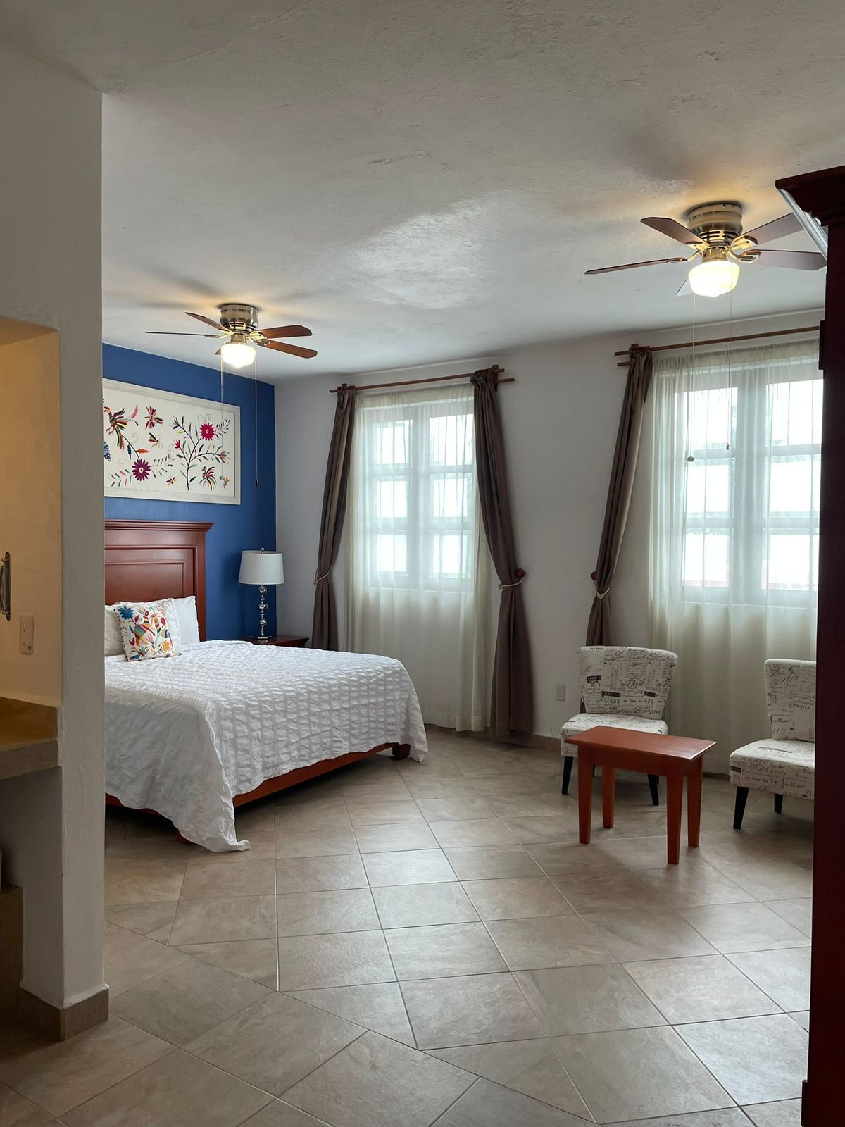 Un dormitorio con una cama, dos sillas, una mesa, un ventilador de techo y un ventanal con cortinas.