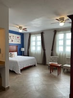 A bedroom with a bed, two chairs, a table, a ceiling fan, and a window with curtains.