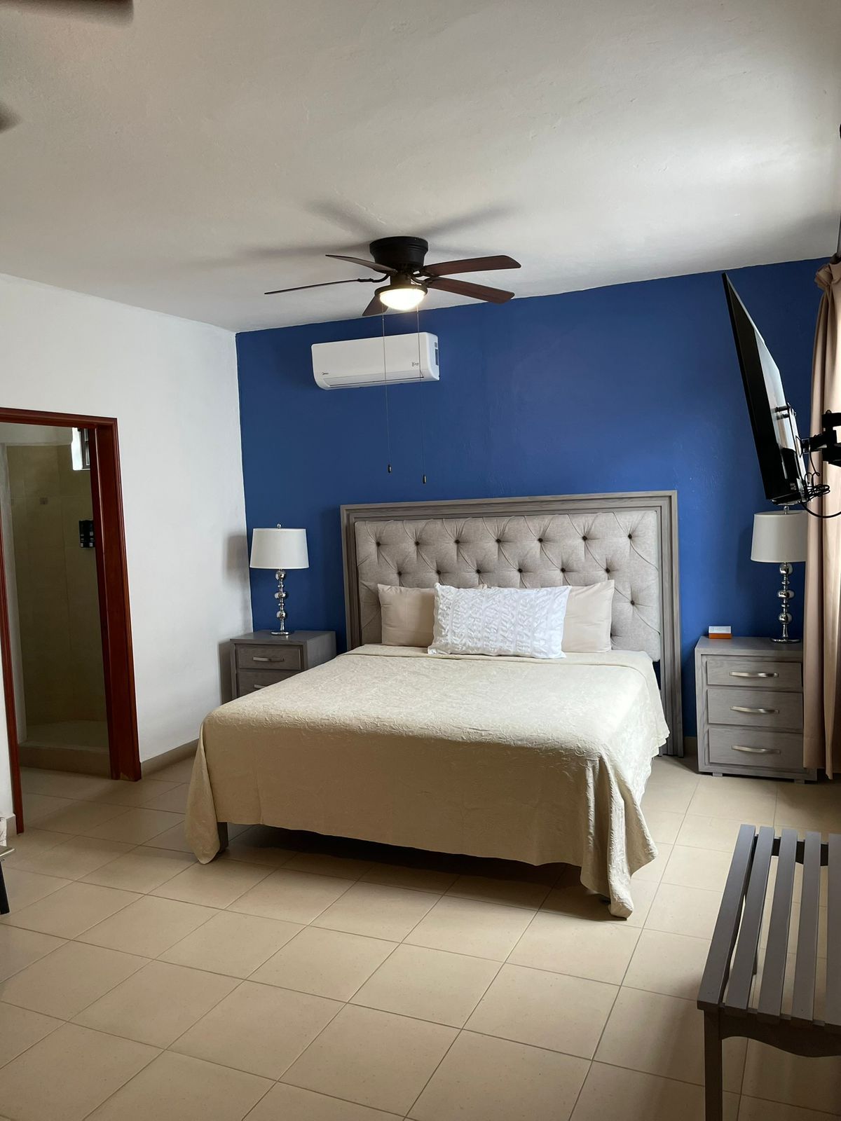 Ein Schlafzimmer mit einem Bett, einem Deckenventilator, einer Klimaanlage, Nachttischen mit Lampen und einer hölzernen Bank.