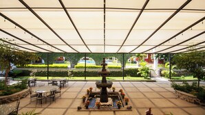 Um pátio exterior com uma fonte central, rodeado por mesas e cadeiras, e vista para um jardim ajardinado através de grandes janelas.