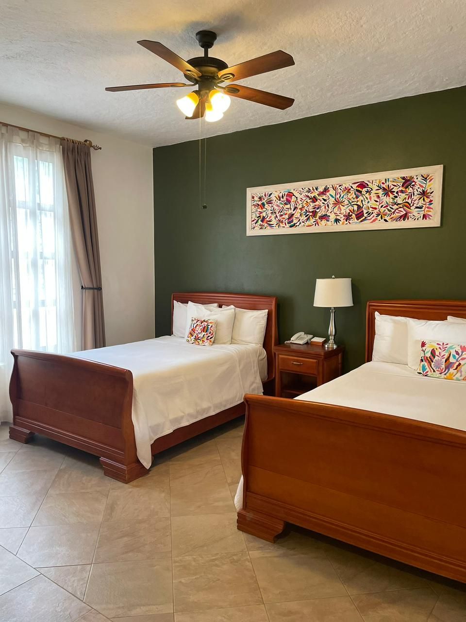 A hotel room with two wooden beds, a ceiling fan, a nightstand with a lamp, and a painting on the wall.