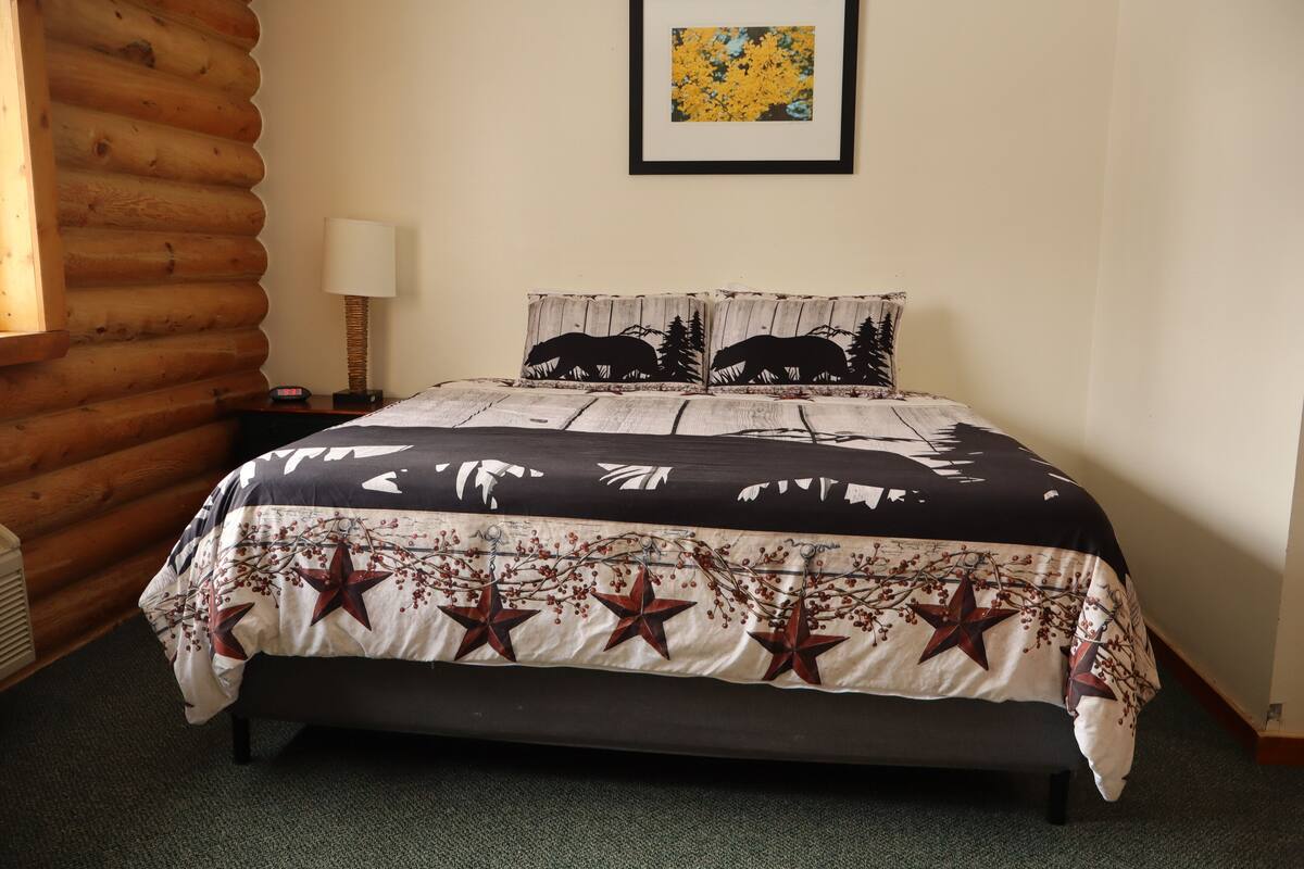 A bed with a star-patterned bedspread, two pillows with a forest design, a framed picture on the wall, and a wooden log wall.