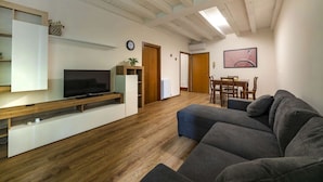 Ein modernes Wohnzimmer mit Holzboden, einer grauen Couch, einem Fernseher auf einem weißen Schrank, einem Esstisch mit Stühlen und einer Uhr an der Wand.