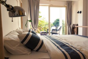 Un letto rifatto con lenzuola a righe, un tavolino con una lampada e una sedia davanti a un balcone con tende.