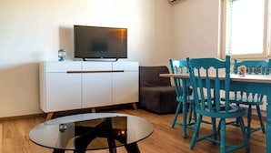 Un salon moderne avec un téléviseur à écran plat posé sur un meuble blanc, une table basse en verre, des fauteuils de couleur turquoise et un canapé gris.