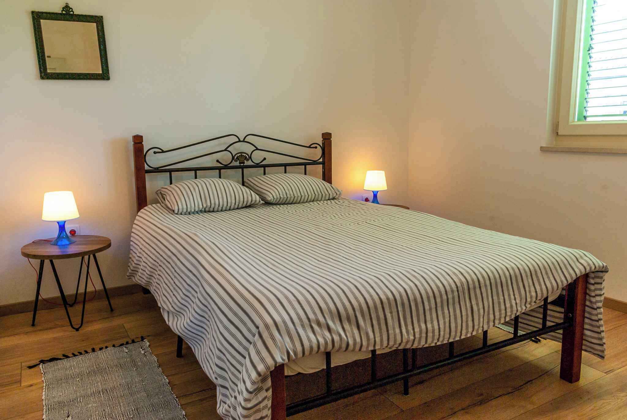 A single bed with a striped bedspread, two bedside lamps, a small rug, and a window with blinds.