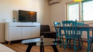 Un salon moderne doté d’un téléviseur à écran plat, d’une table basse en verre, d’une table à manger turquoise avec des chaises, d’un canapé et d’une fenêtre avec des stores.