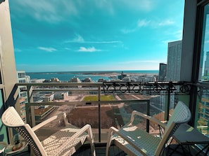 Un balcon avec des fauteuils et une table en osier, offrant une vue sur un paysage urbain avec des bâtiments et une étendue d’eau.