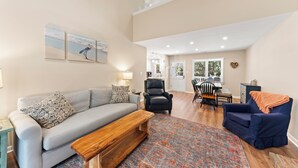 A spacious living room with a grey sofa, a blue armchair, and a wooden coffee table. There is a patterned rug on the floor, and a dining area with a table and chairs is visible in the background.
