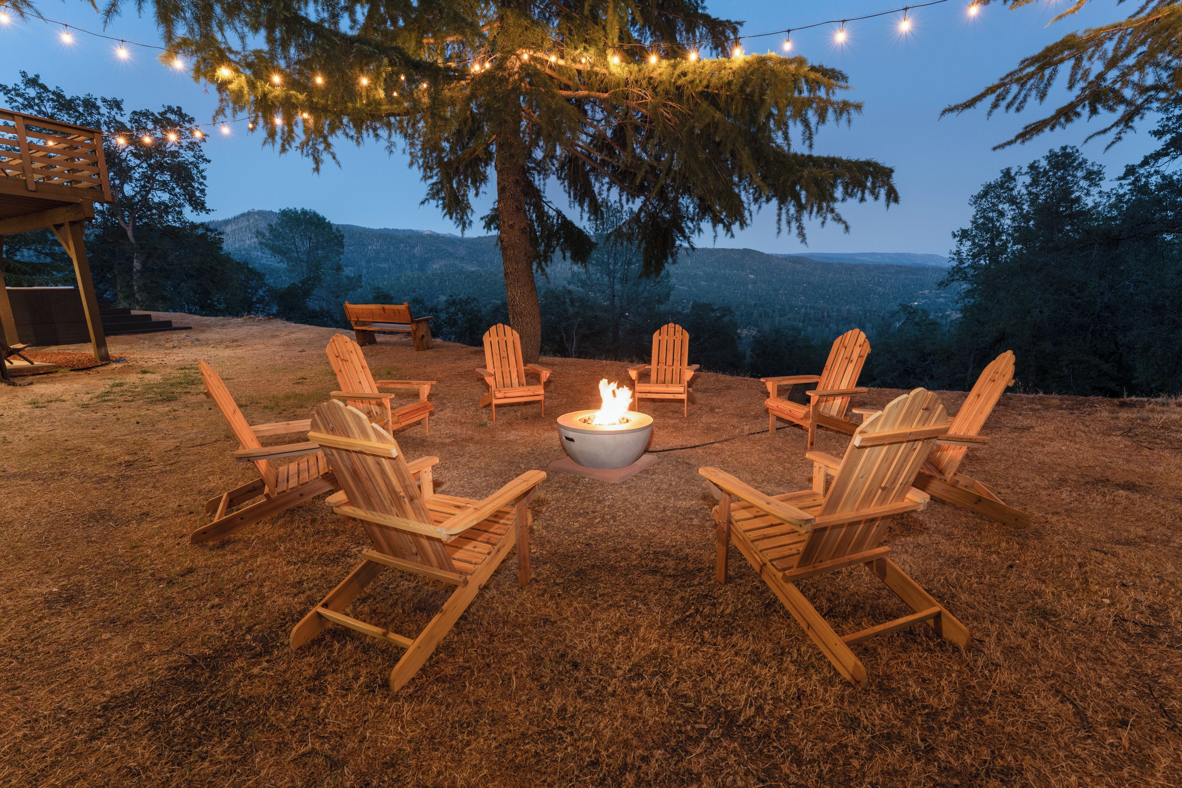 A fire pit surrounded by wooden Adirondack chairs in a grassy outdoor area with string lights overhead.