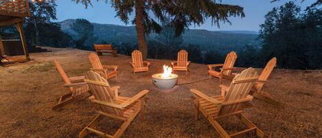 A fire pit surrounded by wooden Adirondack chairs in a grassy outdoor area with string lights overhead.