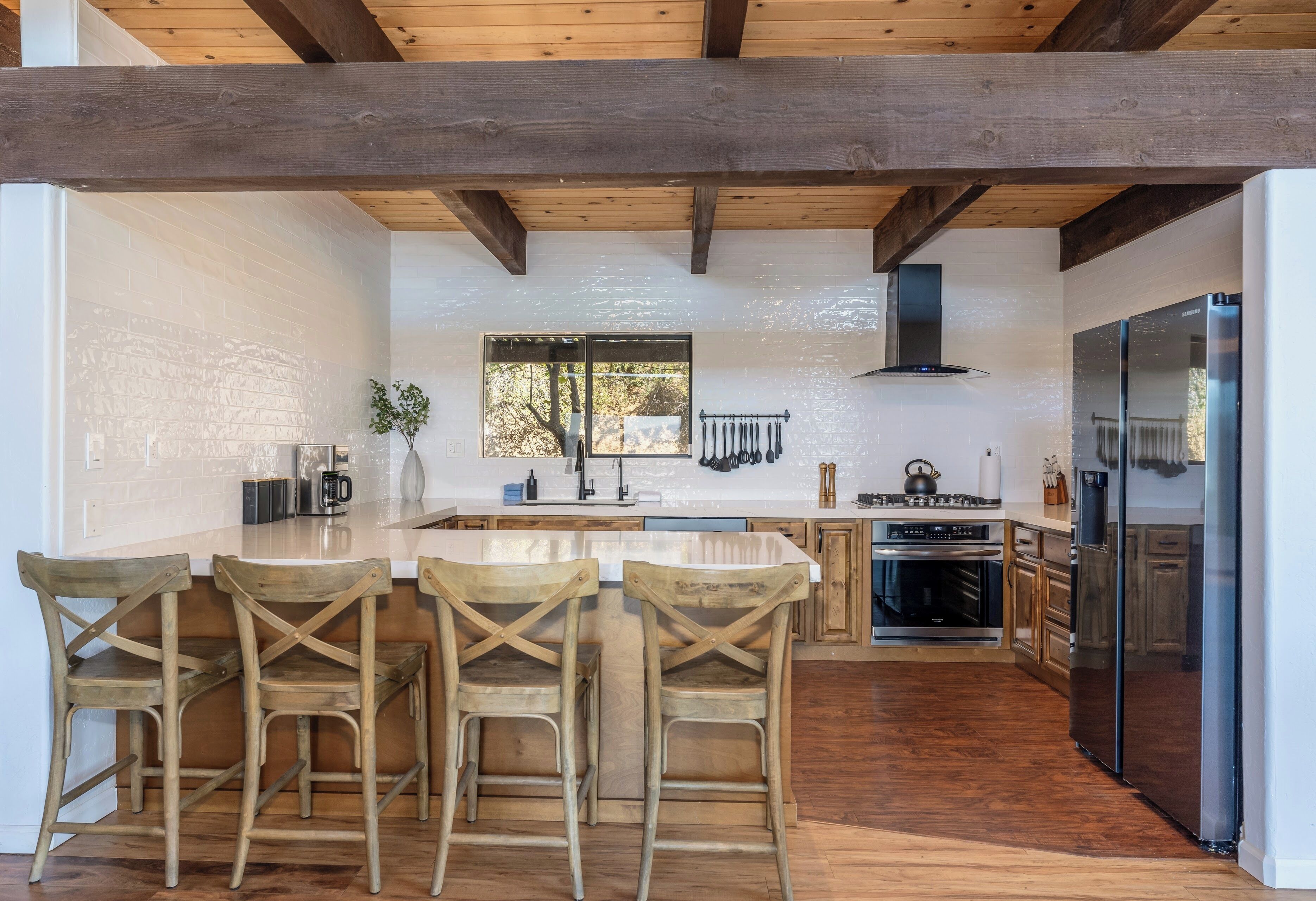 A kitchen with wooden beams, a large island with bar stools, a stainless steel refrigerator, and a window with a view of trees.