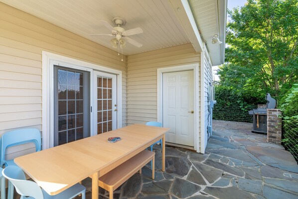 Une terrasse couverte avec une table et des chaises en bois, un ventilateur de plafond et un chemin de pierre menant à un espace barbecue.