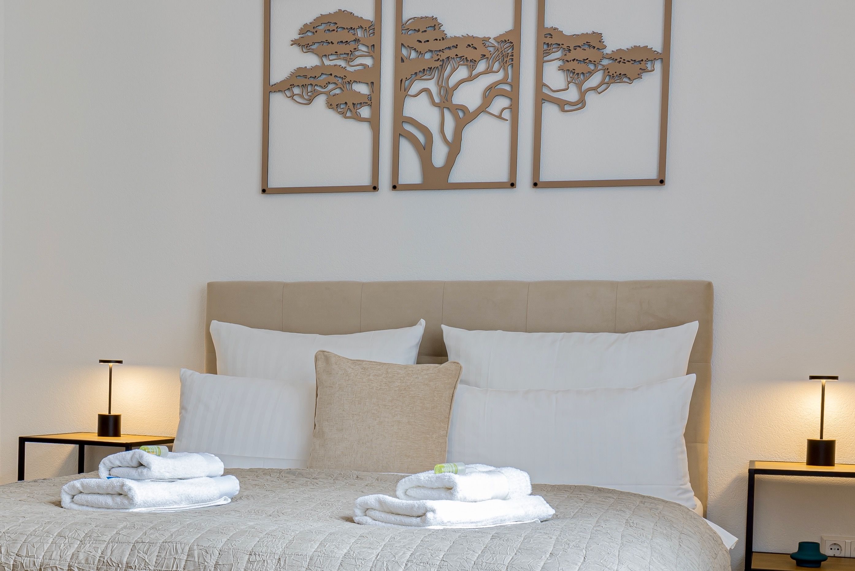Een keurig opgemaakt bed met witte lakens en handdoeken, een beige bekleden hoofdbord, en twee wandlampen.