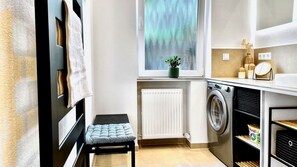 Une petite buanderie comprenant un lave-linge, un radiateur, un petit tabouret et une fenêtre avec une plante.