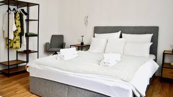 Una habitación de hotel con una cama grande, un sillón tapizado en gris, una mesita de noche con una lámpara y un perchero con diferentes prendas.