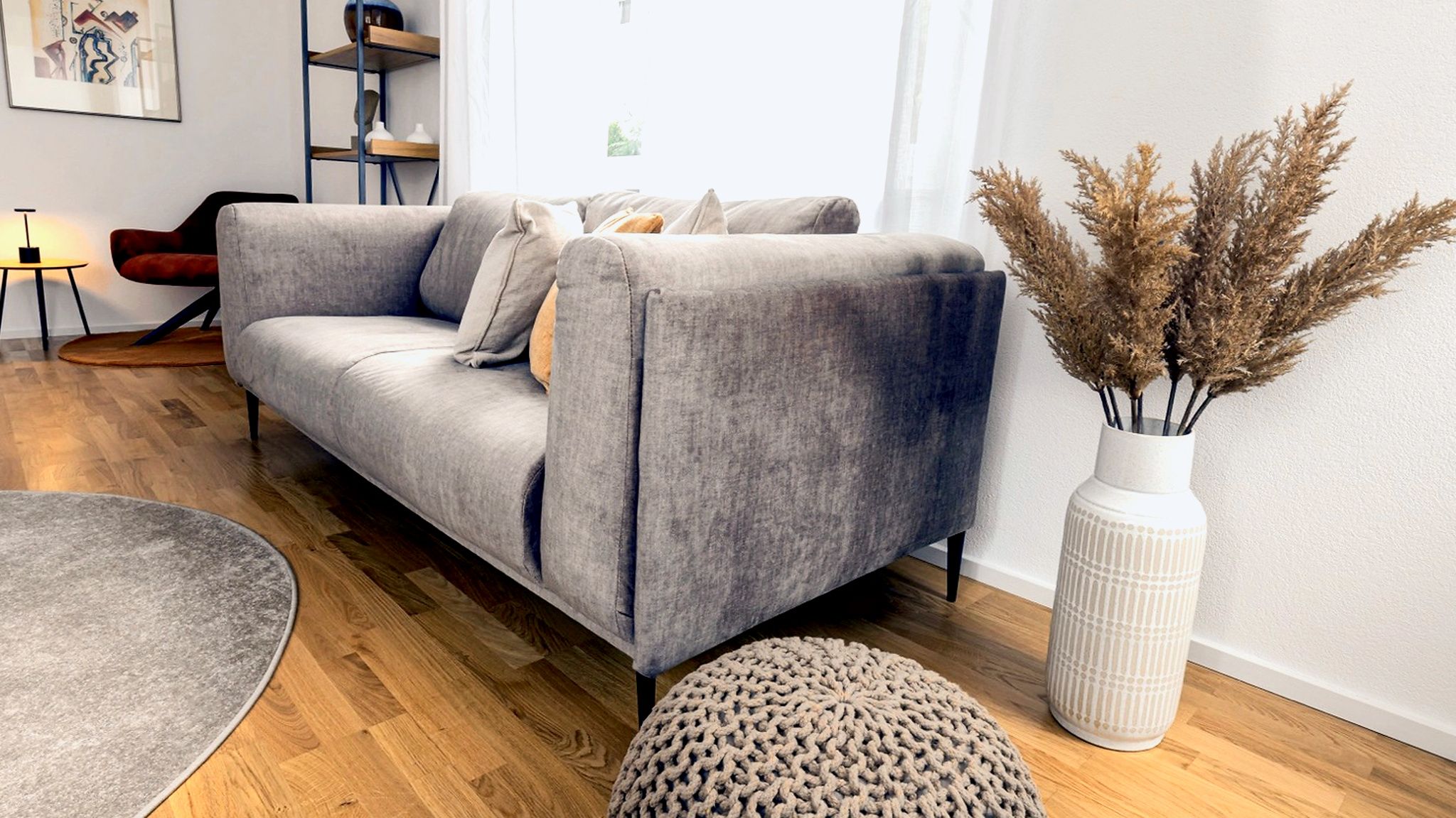 A modern living room with a grey sofa, a round rug, a tall vase with dried plants, and a wooden floor.