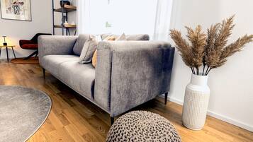 A modern living room with a grey sofa, a round rug, a tall vase with dried plants, and a wooden floor.