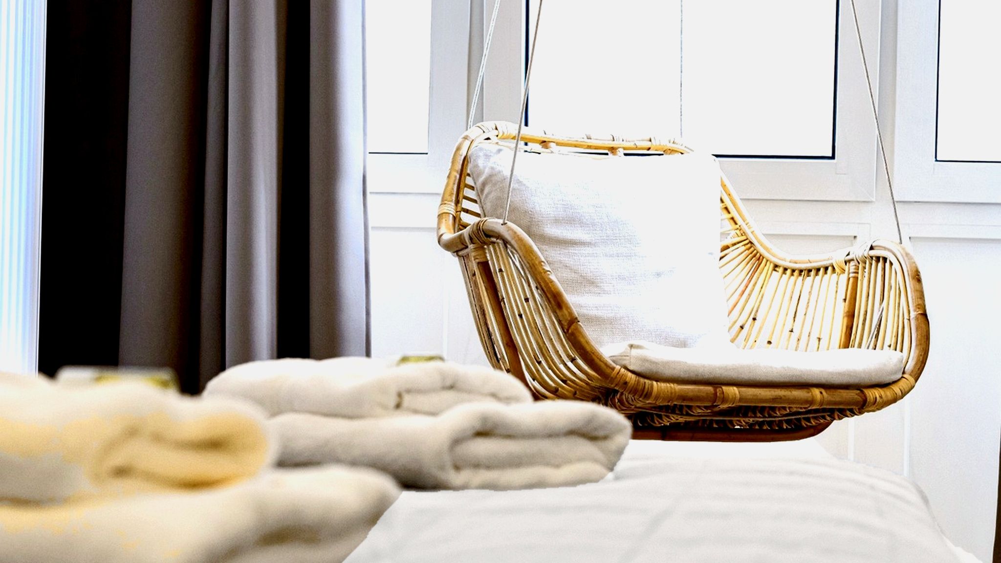 A wicker chair with a cushion, a white bedspread, and a window with curtains in the background.