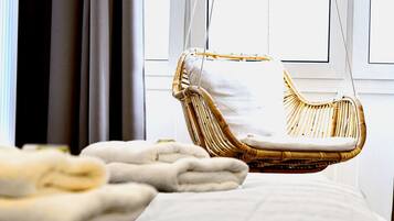 A wicker chair with a cushion, a white bedspread, and a window with curtains in the background.