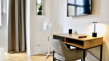 Una habitación moderna con un escritorio de madera, una silla gris, un ventilador de pie, un televisor montado en la pared y una pequeña planta en maceta.