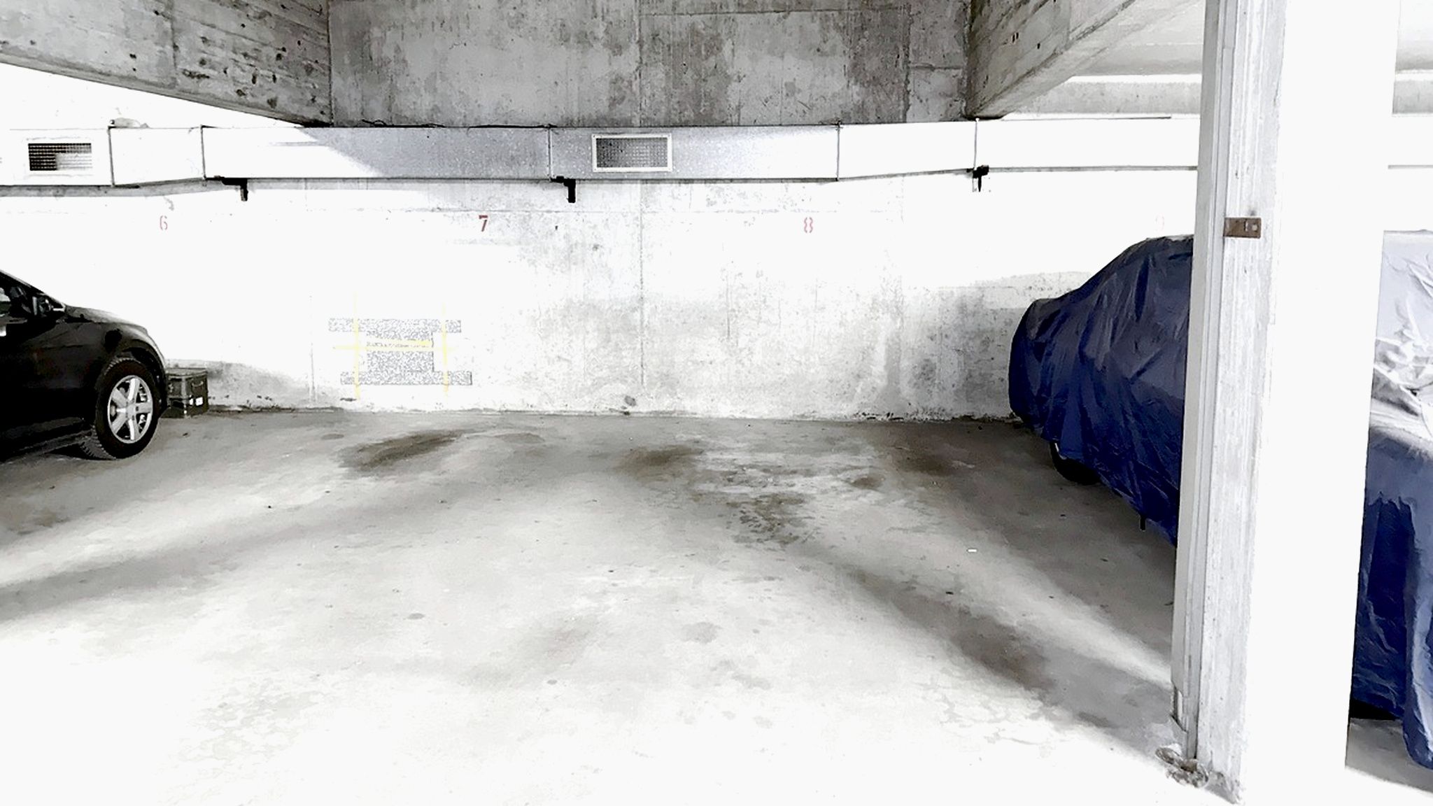 Eine Parkgarage mit Betonboden, ein Auto auf der linken Seite und eine blaue Plane, die ein Objekt auf der rechten Seite bedeckt.