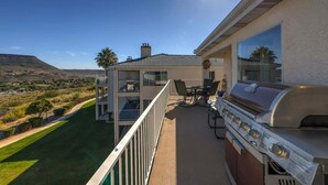 Un balcón con una parrilla y mobiliario exterior, con vista a una zona ajardinada y un edificio de varios pisos.
