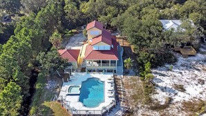 Uma casa grande com piscina, rodeada por árvores e uma pequena área de praia.