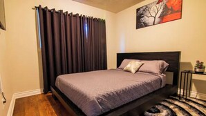 Een slaapkamer met een bed, donkere gordijnen, een schilderij, een klein tafeltje met een plant en een tapijt met een patroon.