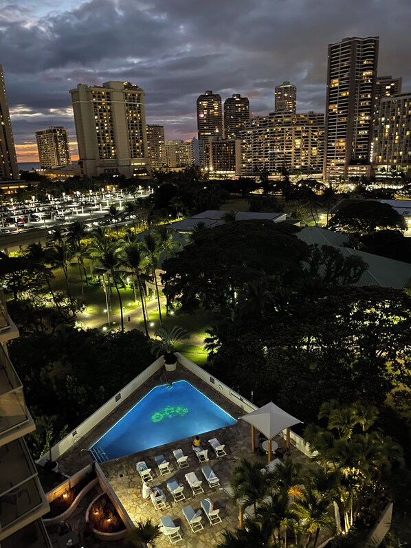 A rooftop pool area with lounge chairs, surrounded by palm trees and a cityscape in the background.