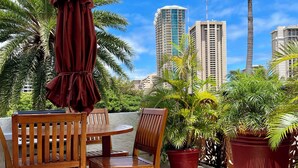 A patio area with wooden chairs and a table, surrounded by potted plants and palm trees, with tall buildings in the background.