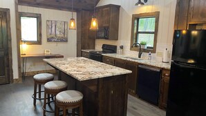 A kitchen with wooden cabinets, a granite countertop, a black refrigerator, and a window with a view of trees. - Whippoorwill Run Cabin — Jasper, AR (Jasper)