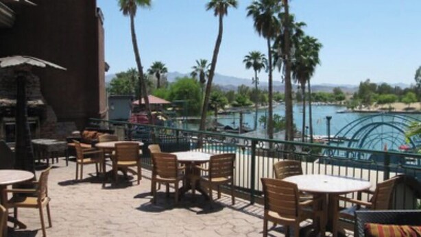 An outdoor patio with wooden tables and chairs, overlooking a pool and palm trees.