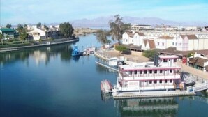 A riverfront hotel with a docked boat, buildings along the river, and a mountainous backdrop.
