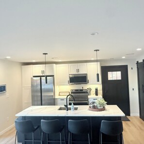 A modern kitchen with a central island, bar stools, stainless steel appliances, and recessed ceiling lighting.