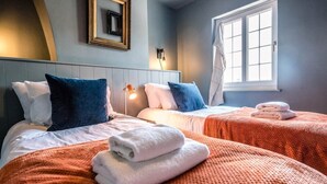 A hotel room with two beds, each with a white and blue pillow, a red blanket, and a folded white towel on the bed. There is a window with a view of the outside, a wall-mounted lamp, and a framed picture on the wall. - Key Cottage, Orford (Orford)