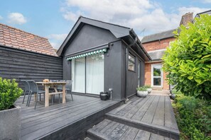 A modern house with a dark exterior, a wooden deck, a dining area with chairs, and a large window.