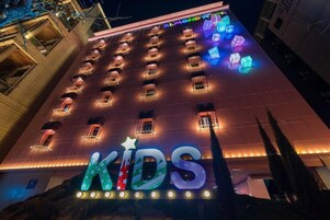 Un hotel con un gran cartel iluminado que dice 'KIDS' en la entrada, varios balcones y un diseño arquitectónico moderno.