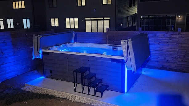 A hot tub with a wooden deck and steps, surrounded by a brick wall and a building with illuminated windows in the background. - PARADISE VILLA 8 (Bowmanville)