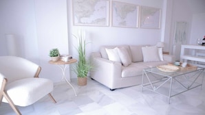A modern living room with a white sofa, a white armchair, a glass coffee table, and a small side table with a plant.