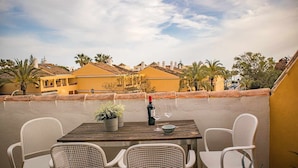 A patio with a wooden table and white wicker chairs, overlooking yellow buildings and palm trees.